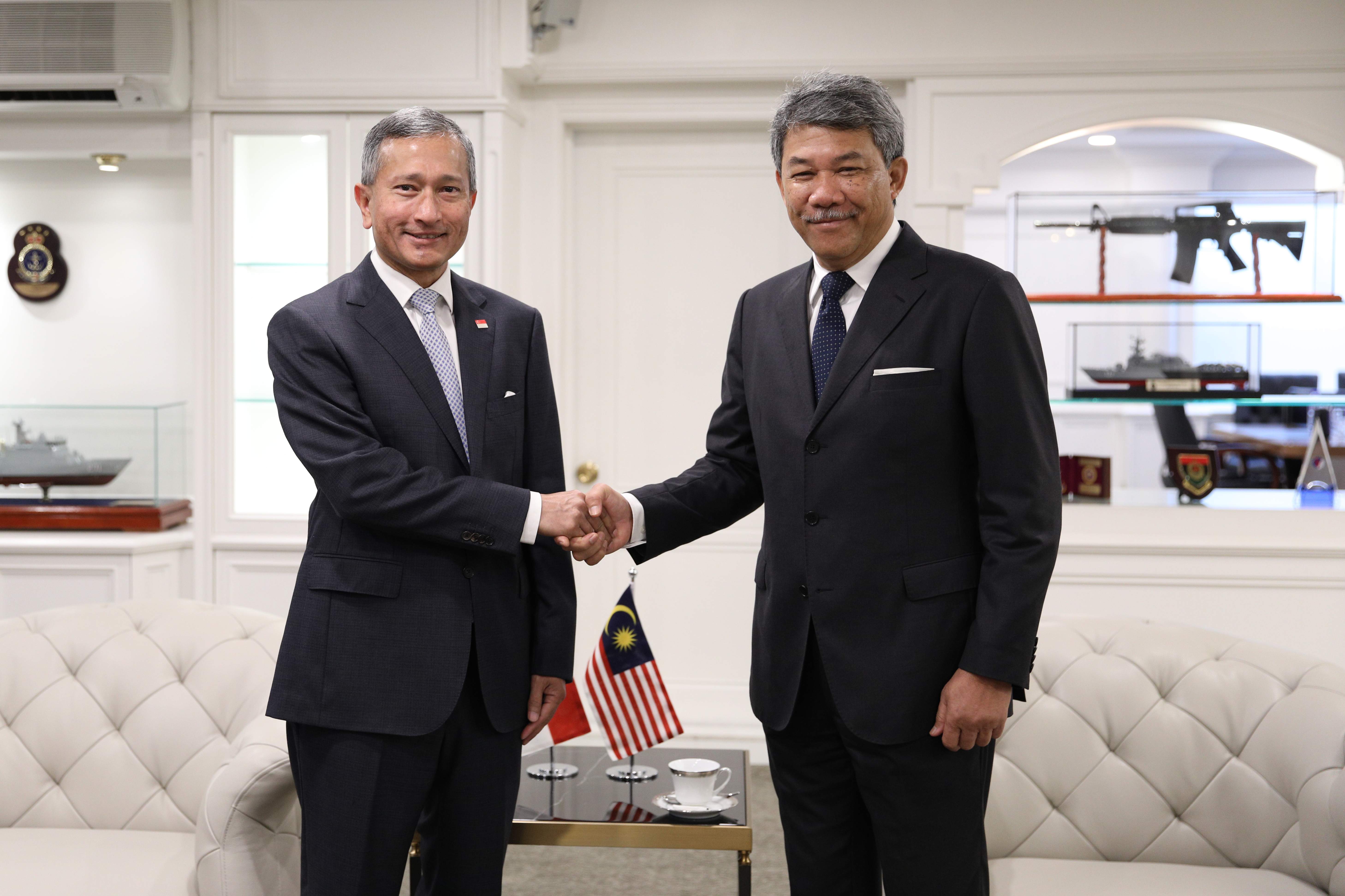 Two men in suits shaking hands, with Singapore and Malaysia flags on a table between them.